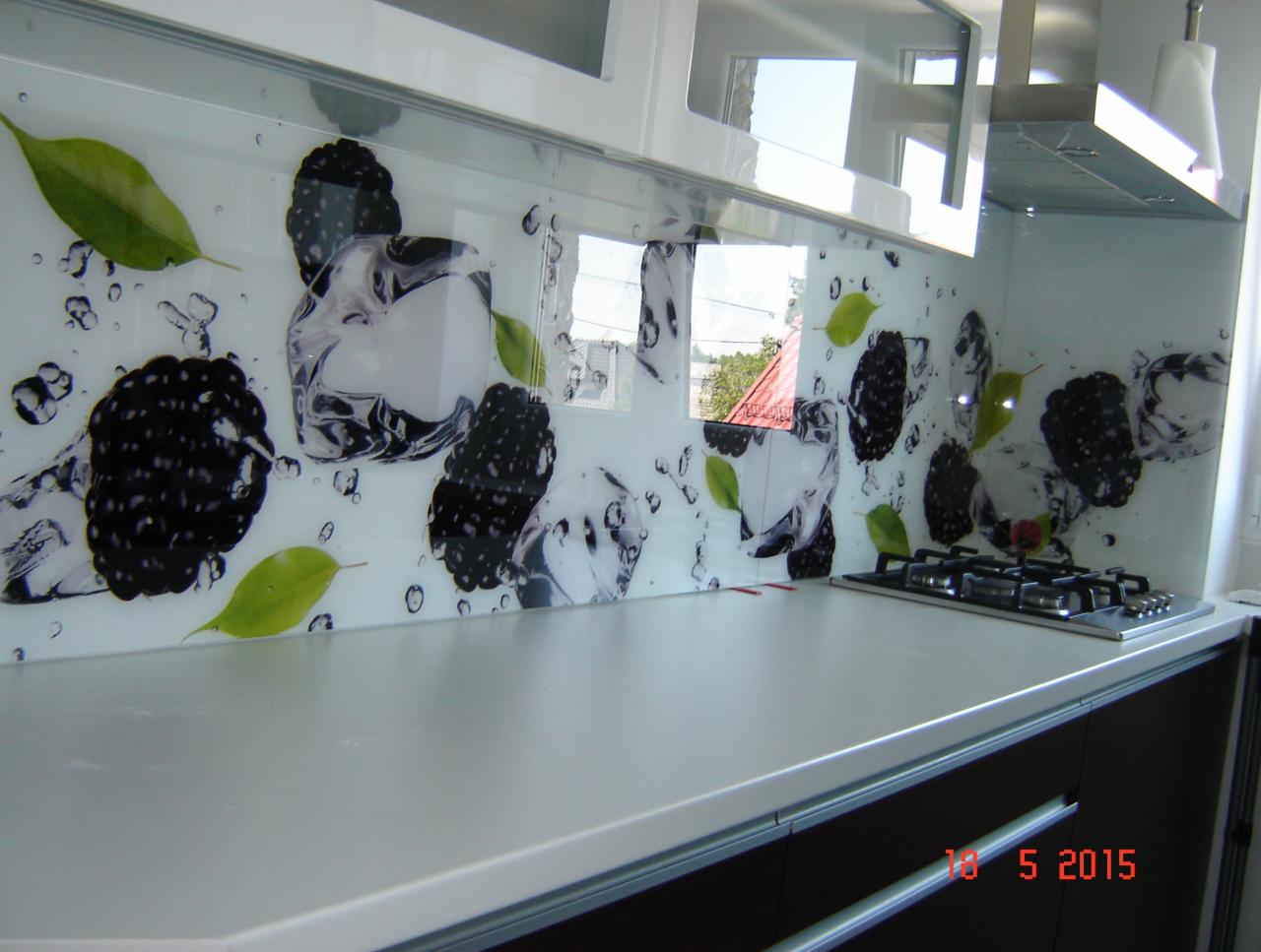 Szklany panel kuchenny z motywem jeżyn, kostek lodu i zielonych liści, zamontowany nad blatem i kuchenką gazową, odbijający światło z okna.