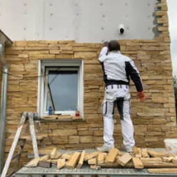 Pracownik w kombinezonie montuje okładzinę z piaskowca na elewacji domu, widoczna drabina i fragmenty okładziny na podeście, częściowo ocieplona ściana z widocznymi mocowaniami, okno i rynna.