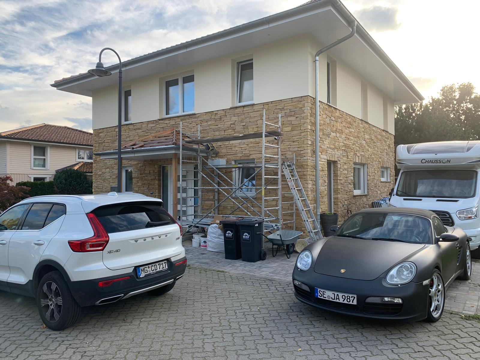 Dwupiętrowy dom z elewacją z kamienia naturalnego w trakcie wykańczania, widoczne rusztowania, drabina, worki z zaprawą, taczka, na podjeździe zaparkowane dwa samochody osobowe i kamper.