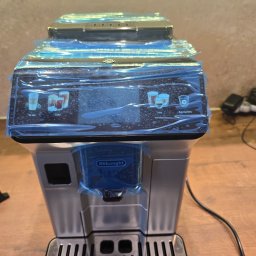 JaroHer Serwis - Nowy ekspres DeLonghi Eletta Explore w srebrnym wykończeniu, z niebieską folią ochronną na dotykowym ekranie z opcjami 'To Go', 'Cold', 'Hot'.
