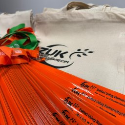 Gadżety i nie tylko - Jasnopomarańczowe wachlarze z czarnym logo ZUK, ułożone wachlarzowo, obok zielono-pomarańczowych smyczy i beżowych toreb z nadrukiem firmowym.