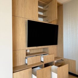 Faktoria Meble - Nowoczesna zabudowa meblowa z jasnego drewna o wyraźnym usłojeniu, z wbudowanym telewizorem, otwartymi szafkami i szufladami z białym wnętrzem, na tle tapety w pionowe paski.