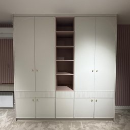 Faktoria Meble - Beżowa szafa wnękowa z subtelnymi łukami na frontach, centralnymi różowymi półkami i ryflowanymi detalami, z eleganckimi złotymi uchwytami.