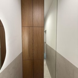 Faktoria Meble - Wysoka, wbudowana szafka z sześcioma drewnianymi panelami, zintegrowana ze ścianą obok lustra. Ściana wykończona szarymi płytkami i białą farbą. Nad szafką widoczna kratka wentylacyjna.