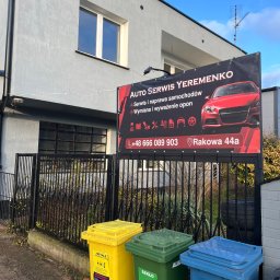 Auto Serwis Yeremenko - Czarny baner 'AUTO SERWIS YEREMENKO' z czerwonym samochodem, zawieszony na metalowym ogrodzeniu przed szarym budynkiem. Na pierwszym planie trzy kolorowe pojemniki do segregacji odpadów.