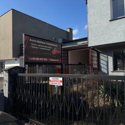 Auto Serwis Yeremenko - Zewnętrzny widok na budynek serwisu samochodowego z dużym czarnym szyldem 'AUTO SERWIS YEREMENKO' reklamującym naprawy i wymianę opon, widoczny za czarnym metalowym ogrodzeniem.