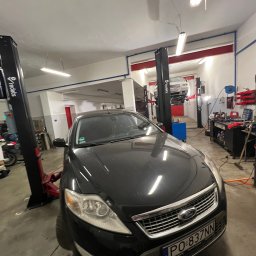Auto Serwis Yeremenko - Czarne Ford Mondeo z polską rejestracją PO 837NN w nowoczesnym warsztacie samochodowym. W tle biały samochód uniesiony na podnośniku Redats, obok stanowisko z narzędziami.