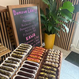 Eklerowski - Ekspozycja domowych eklerów w różnych smakach (czekoladowe, malinowe, pistacjowe) i menu kaw na tablicy, na tle drewnianej ściany i zielonej rośliny.