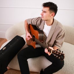 Studio Przystań Piękna Justyna Górecka - Młody mężczyzna w beżowej marynarce gra na brązowej gitarze akustycznej, siedząc na jasnej kanapie bouclé. Obok czarny futerał, w tle biała ściana z ozdobnymi listwami. Spogląda w lewo.