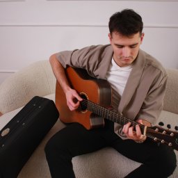 Studio Przystań Piękna Justyna Górecka - Młody mężczyzna w beżowej marynarce gra na drewnianej gitarze akustycznej, siedząc na jasnej, teksturowanej sofie, skupiony na strunach. W tle biała ściana ze sztukaterią, obok czarny futerał.