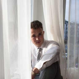Studio Przystań Piękna Justyna Górecka - Młody mężczyzna w białej koszuli i szarym krawacie siedzi na parapecie okna, częściowo ukryty za białymi, półprzezroczystymi firankami, z poważnym wyrazem twarzy.