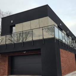 ZYGLASTEEL - Nowoczesny dom z czarną elewacją i ceglanymi wstawkami, z balkonem zabezpieczonym szklanymi panelami na czarnych słupkach, nad ciemną bramą garażową.