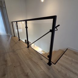 ZYGLASTEEL - Nowoczesne schody z jasnego drewna, z minimalistyczną balustradą ze szkła i czarnych, kwadratowych profili metalowych. Na górze podłoga panelowa, w tle otwarte drzwi.