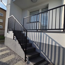 ZYGLASTEEL - Nowoczesne zewnętrzne schody z ciemnymi stopniami i czarną, minimalistyczną balustradą metalową, zamontowaną przy białej, fakturowanej elewacji. Obok ścieżka z jasnego żwiru i betonowych płyt.