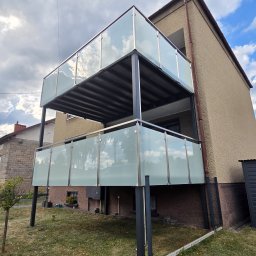 ZYGLASTEEL - Dwupoziomowy balkon z matowymi szklanymi panelami odbijającymi chmury, wsparty na ciemnej metalowej konstrukcji przy beżowym budynku.