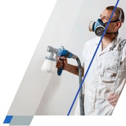 prace-wysokościowe-gorzow.pl - Czyszczenie, mycie, malowanie (natryskowe i ciśnieniowe) na wysokości - Malarz w zabrudzonym kombinezonie i niebieskiej masce ochronnej, trzymający pistolet natryskowy, maluje białą ścianę, patrząc w górę.