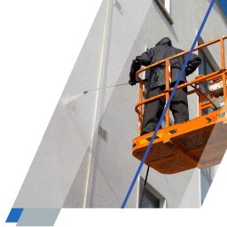 prace-wysokościowe-gorzow.pl - Czyszczenie, mycie, malowanie (natryskowe i ciśnieniowe) na wysokości - Pracownik w ciemnej odzieży ochronnej na pomarańczowym podnośniku koszowym czyści jasną, teksturowaną elewację budynku strumieniem wody pod ciśnieniem.