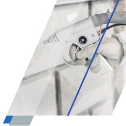 prace-wysokościowe-gorzow.pl - Czyszczenie, mycie, malowanie (natryskowe i ciśnieniowe) na wysokości - Ręka w białej rękawicy i kombinezonie ochronnym trzyma pistolet natryskowy, kierując go na jasną, teksturowaną ścianę, prawdopodobnie podczas malowania lub czyszczenia elewacji budynku.