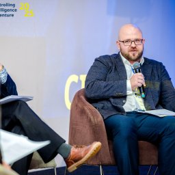 EcoPan - Prelegent w okularach i wzorzystej marynarce, z mikrofonem w dłoni, przemawia podczas panelu dyskusyjnego na tle brandingowym 'CIA controlling intelligence adventure 20 25'.