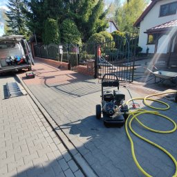 EXPLOTER - Profesjonalna myjka ciśnieniowa z żółtymi wężami na szarej kostce brukowej przed domem, obok otwarty bus serwisowy. W tle osoba czyści nawierzchnię.
