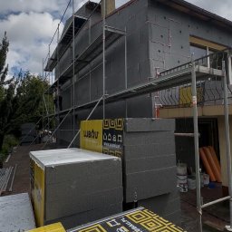KJR Usługi Budowlane - Budynek mieszkalny w trakcie ocieplania ciemnoszarym styropianem grafitowym, z rusztowaniem i pracownikiem. Widoczne stosy płyt izolacyjnych Lubau na pierwszym planie.
