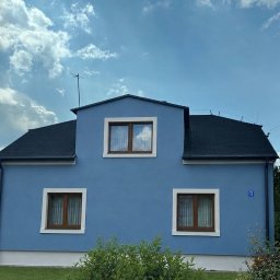 KJR Usługi Budowlane - Niebieski dom jednorodzinny z ciemnym dachem i białymi obramowaniami okien, otoczony zielenią, pod częściowo zachmurzonym niebem. Widoczny numer domu '13'.