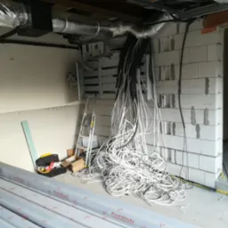 Wnętrze w trakcie prac remontowych: widoczna rozdzielnica elektryczna, pęki kabli, drabina, narzędzia i materiały budowlane, surowy stan ścian.