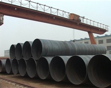 Stos rur stalowych o dużej średnicy, ułożonych na zewnątrz, z widocznym żurawiem w tle.