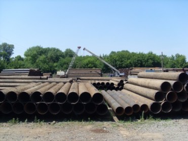 Stos rur stalowych z widocznymi śladami korozji, ułożonych na placu składowym, w tle drzewa i żuraw samochodowy.