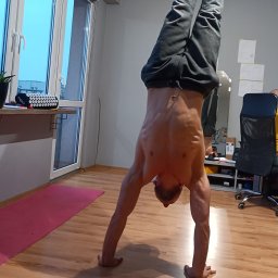 Jakub Pabiańczyk - Muskularny mężczyzna bez koszulki wykonuje precyzyjne stanie na rękach na drewnianej podłodze, obok różowej maty. W tle duże okno i biurowe krzesło.