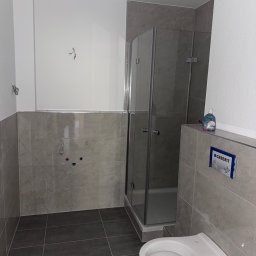 Dawid Pazurek - Nowoczesna łazienka z ciemnoszarą podłogą i jasnoszarymi płytkami ściennymi, białym podwieszanym WC, kabiną prysznicową, panelem Geberit i widocznymi przyłączami wodnymi.