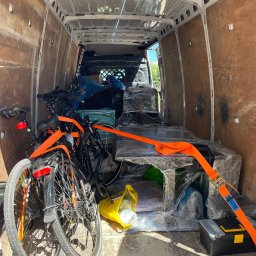 Marcin Wirtel - Wnętrze białego busa transportowego, załadowane dwoma rowerami miejskimi i owiniętym stołem, zabezpieczone pomarańczowymi pasami transportowymi. Widoczne drewniane panele boczne.