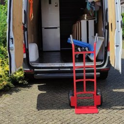 Marcin Wirtel - Biały bus z otwartymi tylnymi drzwiami, częściowo załadowany srebrną lodówką i innymi przedmiotami, z czerwonym wózkiem transportowym na brukowanym podjeździe.