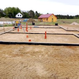 Zakład Remontowo Budowlany DOMEX Marek Krzak - Przygotowane fundamenty domu jednorodzinnego z widocznymi rurami instalacyjnymi i czarną folią kubełkową. Piaszczysty teren budowy, w tle dwa domy i zalesiony krajobraz.