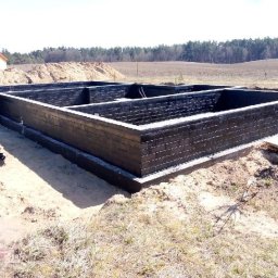 Zakład Remontowo Budowlany DOMEX Marek Krzak - Solidne ściany fundamentowe domu jednorodzinnego, pokryte czarną izolacją przeciwwilgociową, wkopane w piaszczysty grunt na tle odległego lasu i jasnego nieba.