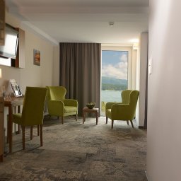 Skalny SPA - Hotel Solina w Bieszczadach Polańczyk - Nowoczesny pokój z dwoma zielonymi fotelami i stolikiem, z dużym oknem na jezioro i góry. W tle biurko z telewizorem, na podłodze wzorzysty dywan.