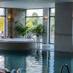 Skalny SPA - Hotel Solina w Bieszczadach Polańczyk - Nowoczesny basen wewnętrzny z okrągłym jacuzzi, w którym kobieta relaksuje się z telefonem. Panoramiczne okna z widokiem na zieleń, kamienne ściany i leżaki na tarasie tworzą luksusową atmosferę.