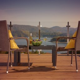 Skalny SPA - Hotel Solina w Bieszczadach Polańczyk - Dwa wiklinowe fotele z żółtymi poduszkami i stolik z winem oraz owocami na drewnianym tarasie z widokiem na jezioro i góry, pod błękitnym niebem.