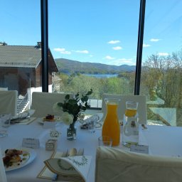 Skalny SPA - Hotel Solina w Bieszczadach Polańczyk - Elegancko zastawiony stół z białymi obrusami, kwiatami i daniami z szynką, z widokiem przez panoramiczne okno na jezioro, góry i dom z brązowym dachem.