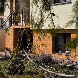Innowacyjny Dom - Srebrna jednostka zewnętrzna z poziomymi lamelami, zamontowana na stelażu przy domu z beżową elewacją i pomarańczowymi płytkami, częściowo zasłonięta rozmywającymi się gałęziami.