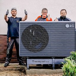 Innowacyjny Dom - Trzech uśmiechniętych instalatorów w roboczych strojach, w tym kobieta, prezentuje dużą, ciemnoszarą jednostkę zewnętrzną LG z widocznym napisem 'INNOWACYJNY Dom', dając kciuki w górę. Urządzenie...
