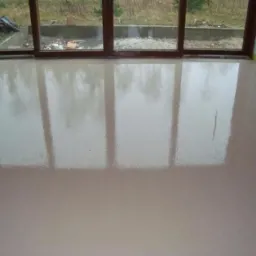 Świeżo wylana, beżowa posadzka jastrychowa w pomieszczeniu z dużym oknem tarasowym, odbijającym krajobraz zewnętrzny. Widoczna drobna rysa na powierzchni posadzki.