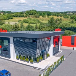 HANDEL TNP - Nowoczesna siedziba firmy Redan z przeszkloną fasadą, poziomymi żaluzjami i doniczkowymi tujami, obok czerwony magazyn, na tle zielonych wzgórz.