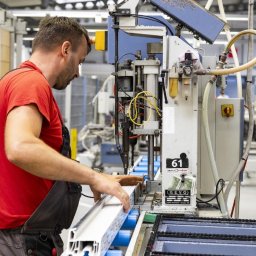 HANDEL TNP - Mężczyzna w czerwonej koszulce precyzyjnie podaje biały profil okienny do maszyny produkcyjnej z etykietą 'okna redan' w fabryce.
