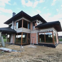 MasterHome - Nowoczesny dom w budowie z dużymi, brązowymi oknami i szklanymi drzwiami tarasowymi, częściowo wykończony ciemnym dachem. Widoczne surowe ściany z cegły i betonu, otoczenie budowlane.