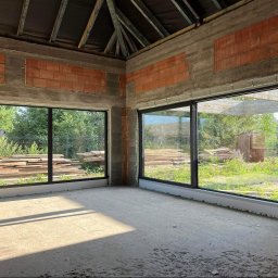 MasterHome - Wnętrze budynku w stanie surowym z dużymi, czarnymi oknami panoramicznymi, przez które widać zieleń i materiały budowlane. Widoczne betonowe ściany, cegły i drewniana konstrukcja dachu.