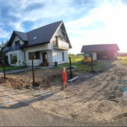 MasterHome - Nowoczesny dom jednorodzinny z ciemnym dachem i jasną elewacją, na posesji z częściowo zamontowanymi słupkami ogrodzenia. Słoneczny dzień, długie cienie, małe dziecko w pomarańczowym ubraniu...