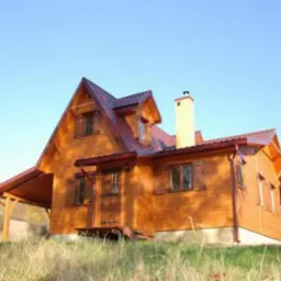 Zewnętrzny widok na nowy, parterowy dom z drewna z poddaszem użytkowym, brązowymi okiennicami i jasnym kominem, otoczony wysoką trawą i drzewami w tle.