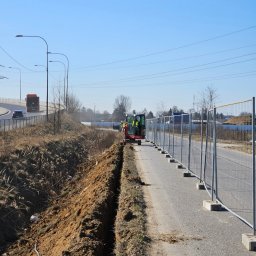 Elektrotechnika Konrad Wykowski - Czerwona mini koparka kopie świeżo wykopany rów wzdłuż asfaltowej ścieżki, obok tymczasowe ogrodzenie. W tle estakada z samochodami, słupy oświetleniowe i błękitne niebo.