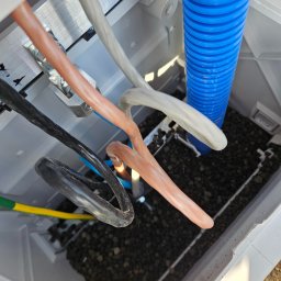 Elektrotechnika Konrad Wykowski - Otwarta skrzynka przyłączeniowa z grubymi kablami (czarny, brązowy, szary) i niebieską rurą karbowaną, osadzonymi w ciemnym kruszywie, widok z góry.